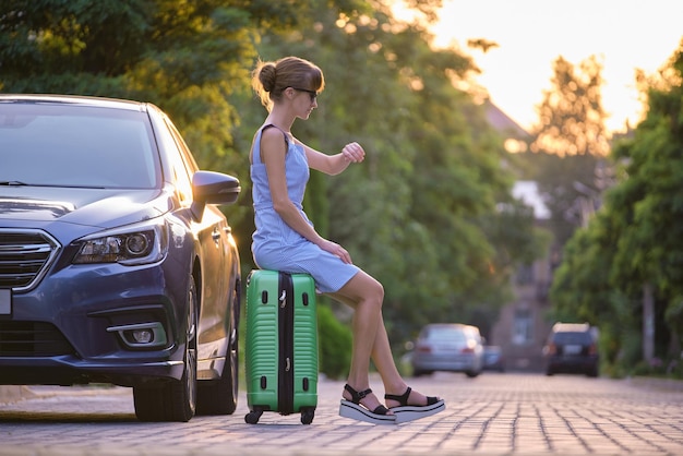 Young tired woman with suitcase sitting near her car waiting for someone Travel and vacations concept