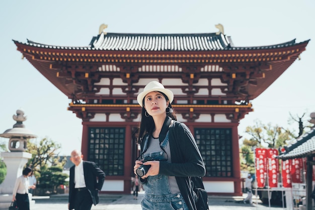 Young tourist backpacker girl with camera traveling in ancient japanese temple on summer holidays in osaka japan. woman photographer shooting in shitennoji on sunny day with professional digicam.