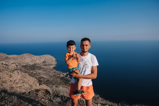 A young tourist with his young son climbed the mountains A man and a boy are standing at the top against the background of the blue sea Active recreation