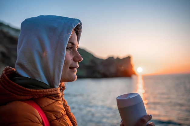 A young tourist woman holding coffee tumbler cup while sitting outdoor and enjoying sunrise over sea