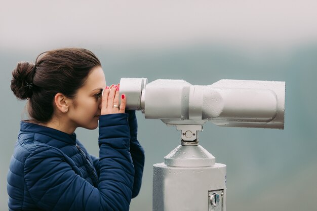 Young tourist woman looking through coin operated high powered binoculars on the Caucasus Mountains, Kazbegi, Georgia