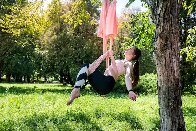 Young trainer with gorgeous hair from fly yoga is engaged in stretching on an air hammock attached to a tree Yoga fly concept on air