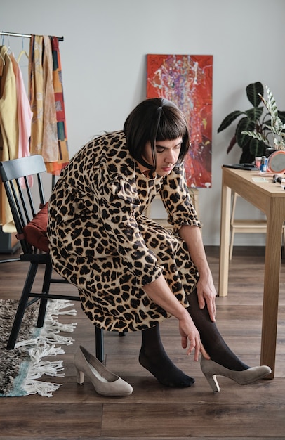 Young transgender in female clothes sitting on chair and putting on shoes in dressing room