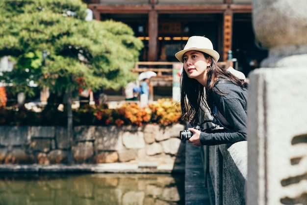 young travel lens man holding slr camera relying on the stone bridge sightseeing osaka outdoor in shitennoji. girl tourist with straw hat standing above the pond with trees beside. trip jp lifestyle.