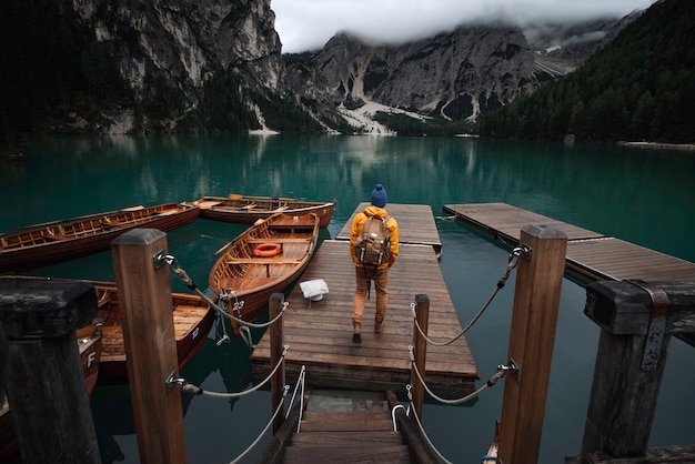 Young traveler boy with blue hat, vintage backpack and yellow raincoat on jetty of lago di Braies surrounded by mountains of Italian alps