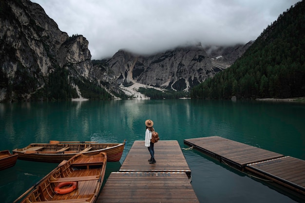 Young traveler girl with hat and vintage backpack on jetty of lago di Braies surrounded by the mountains of the Italian alps