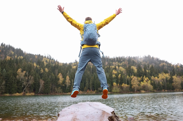 Young traveler jumps against backdrop of forest and lake raising hands to sky