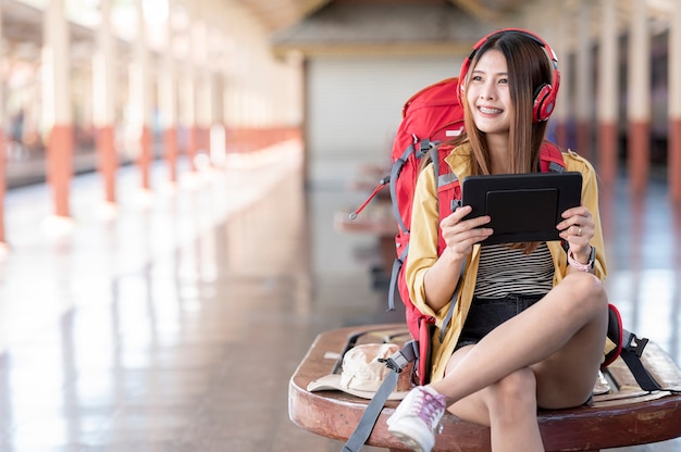 Young traveler woman with backpack and headphone holding tablet while sitting at railway station, waiting for departure