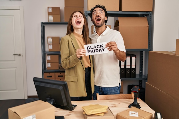 Young two people holding black friday banner at small business store angry and mad screaming frustrated and furious shouting with anger looking up