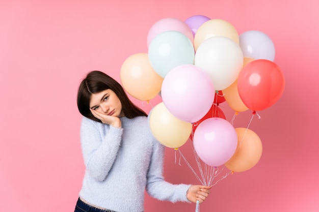 Young Ukrainian teenager girl holding lots of balloons over pink wall unhappy and frustrated