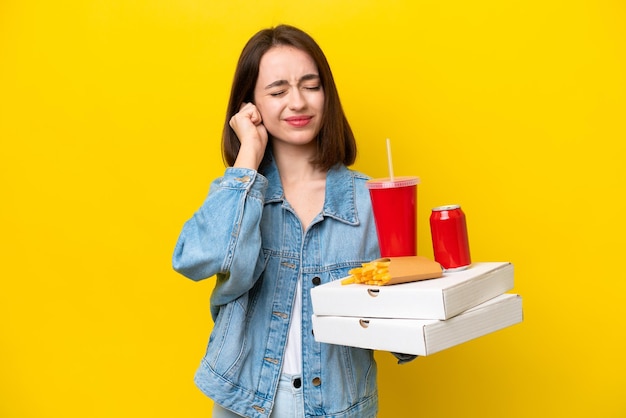 Young Ukrainian woman holding fast food isolated on yellow background frustrated and covering ears