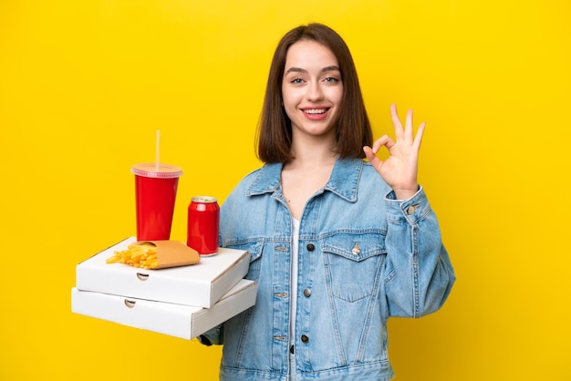 Young Ukrainian woman holding fast food isolated on yellow background showing ok sign with fingers