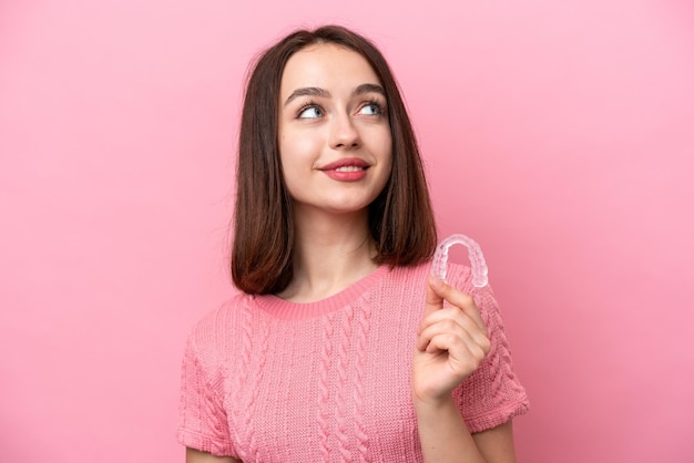 Young Ukrainian woman holding invisible braces isolated on pink background looking up while smiling