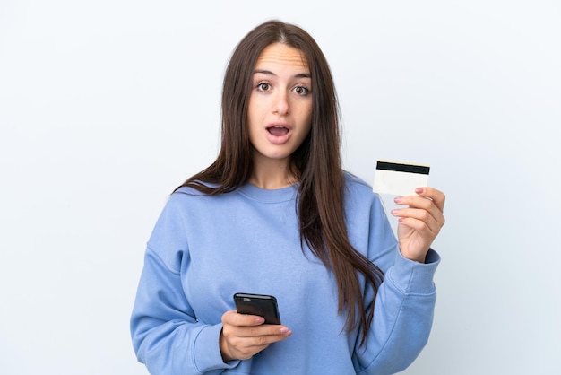 Young Ukrainian woman isolated on white background buying with the mobile and holding a credit card with surprised expression