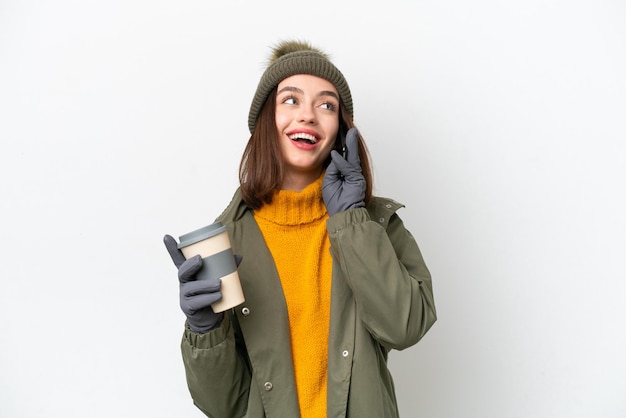 Young Ukrainian woman wearing winter jacket isolated on white background holding coffee to take away and a mobile