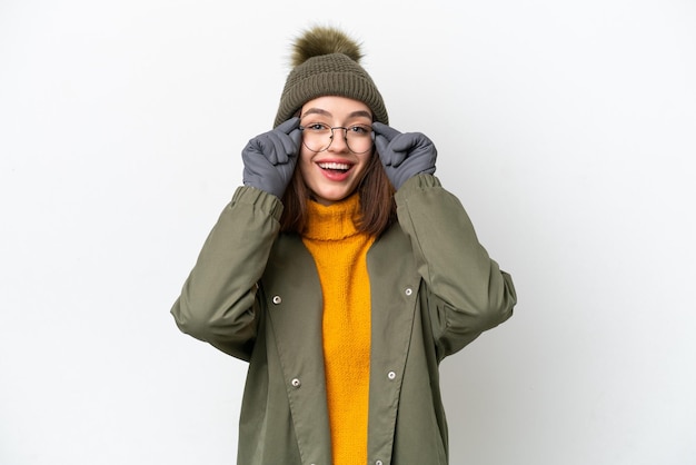 Young Ukrainian woman wearing winter jacket isolated on white background with glasses and surprised