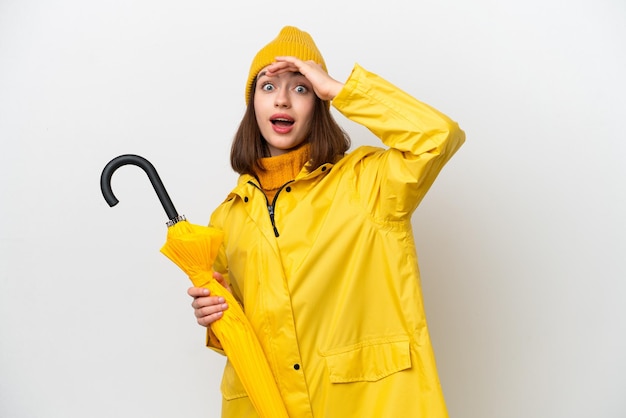 Young Ukrainian woman with rainproof coat and umbrella isolated on white background doing surprise gesture while looking to the side