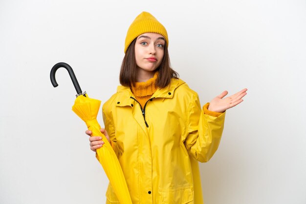 Young Ukrainian woman with rainproof coat and umbrella isolated on white background having doubts while raising hands