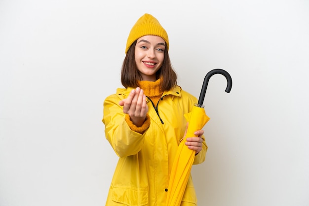 Young Ukrainian woman with rainproof coat and umbrella isolated on white background inviting to come with hand Happy that you came