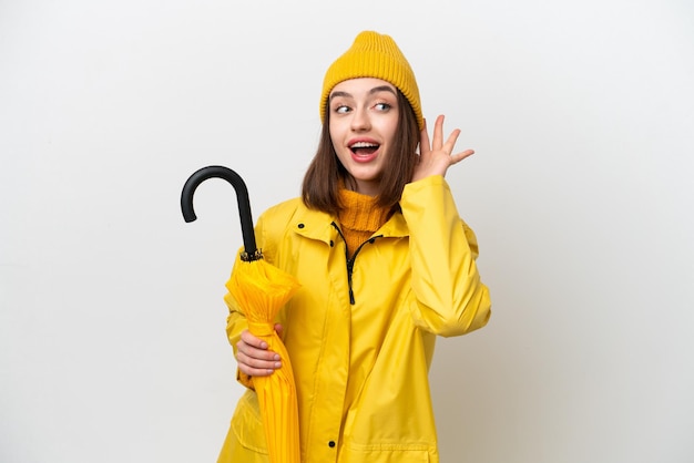 Young Ukrainian woman with rainproof coat and umbrella isolated on white background listening to something by putting hand on the ear