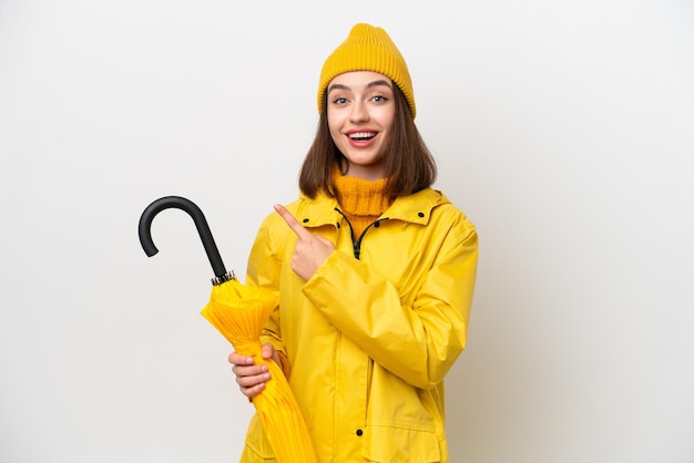 Young Ukrainian woman with rainproof coat and umbrella isolated on white background pointing to the side to present a product