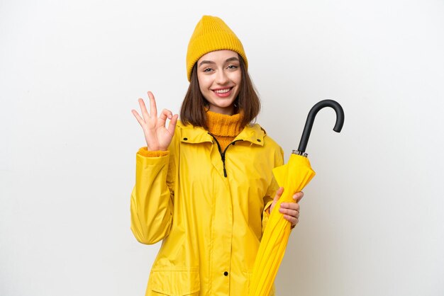 Young Ukrainian woman with rainproof coat and umbrella isolated on white background showing ok sign with fingers