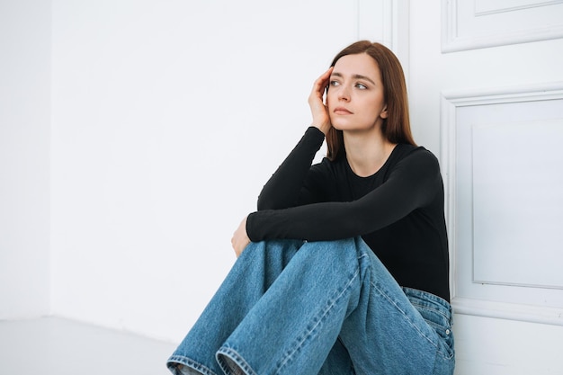 Young unhappy woman teenager girl with long hair in jeans sitting on floor in closed position by door at home negative emotion