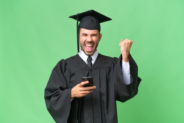 Young university graduate man over isolated background with phone in victory position