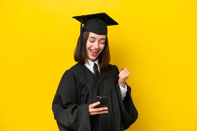 Young university graduate Ukrainian woman isolated on yellow background with phone in victory position