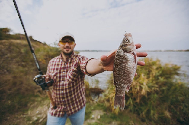 Young unshaven man in checkered shirt, cap, sunglasses holds fishing pole and extends his hand to caught fish on shore of lake near shrubs and reeds. Lifestyle, recreation, fisherman leisure concept.