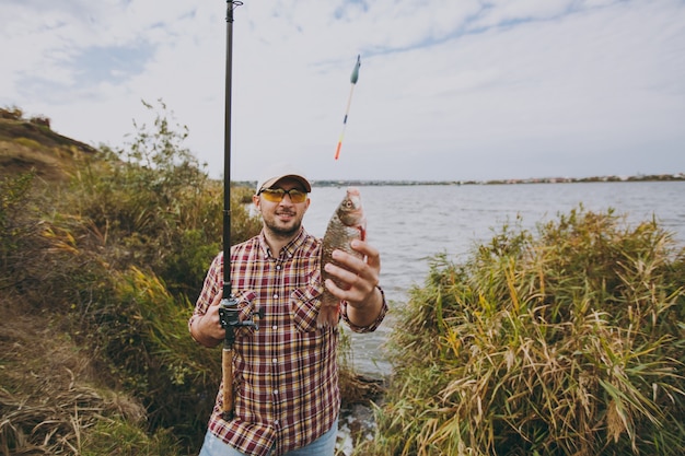 Young unshaven man in checkered shirt, cap, sunglasses holds fishing pole and extends his hand to caught fish on shore of lake near shrubs and reeds. Lifestyle, recreation, fisherman leisure concept.