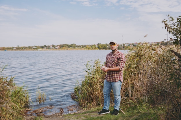 Young unshaven man in checkered shirt, cap and sunglasses stand by lake and holds small box with maggots against background of water, shrubs and reeds. Lifestyle, fisherman recreation, leisure concept