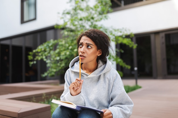 Young upset lady with dark curly hair sitting with notebook on knees and thoughtfully 