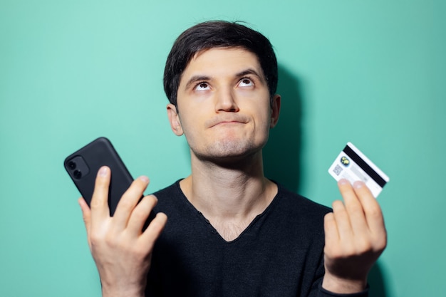 young upset man looking up holding in hands smartphone and credit card on wall of aqua menthe color.