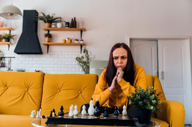 Young upset woman playing chess sitting on sofa Distressed female plays in logical board game with herself in room