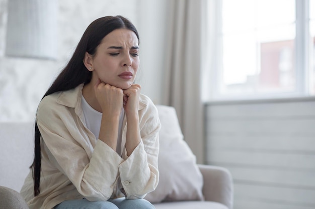 Young upset woman sitting on sofa at home eyes closed hands folded together near face suffers from