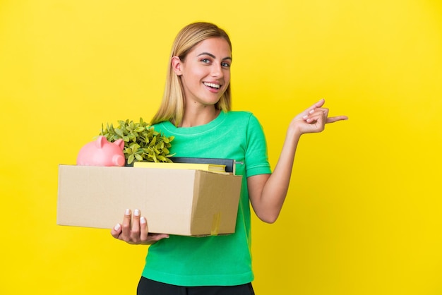 Young Uruguayan girl making a move while picking up a box full of things isolated on yellow background pointing finger to the side