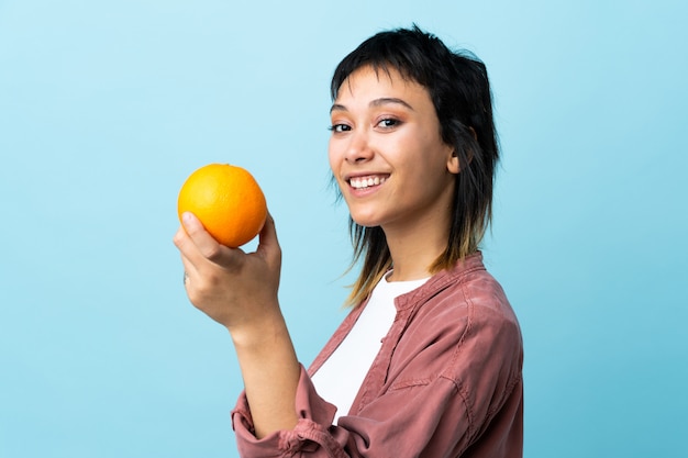 Young Uruguayan woman over blue wall holding an orange