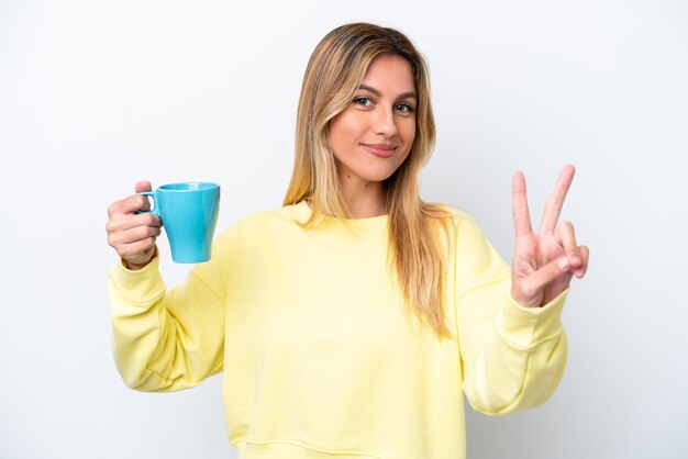 Young Uruguayan woman holding cup of coffee isolated on white background smiling and showing victory sign