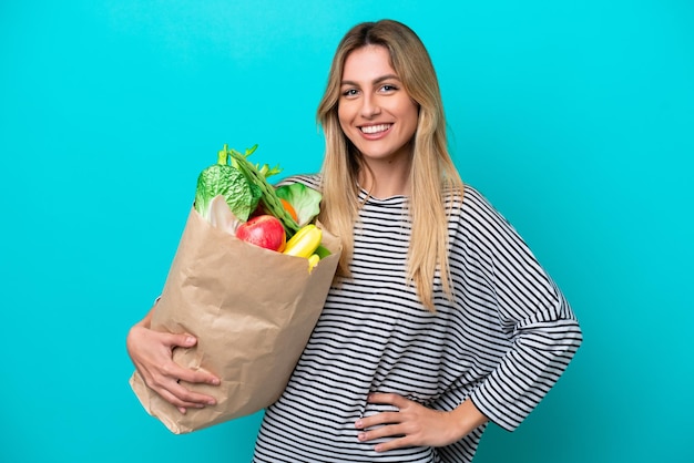 Young Uruguayan woman holding a grocery shopping bag isolated on blue background posing with arms at hip and smiling