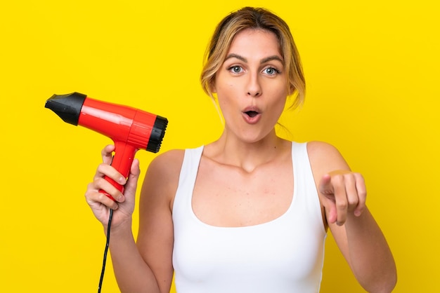 Young Uruguayan woman holding a hairdryer isolate don yellow background surprised and pointing front