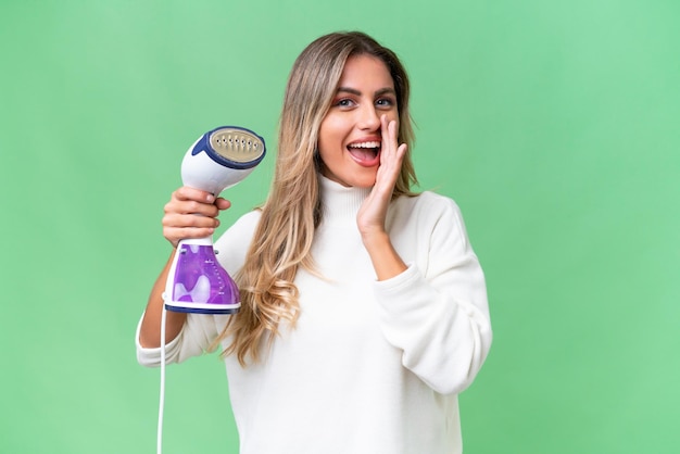 Young Uruguayan woman holding an iron over isolated background shouting with mouth wide open