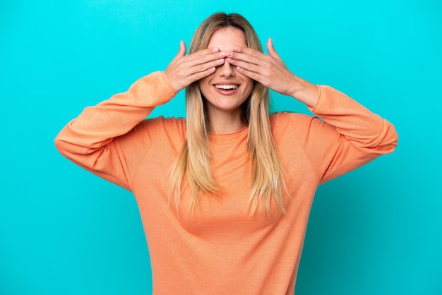 Young Uruguayan woman isolated on blue background covering eyes by hands and smiling