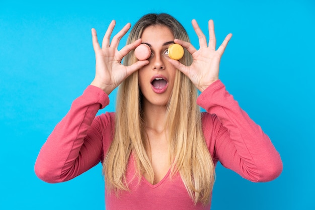 Young Uruguayan woman over isolated blue wall holding colorful French macarons with surprised expression