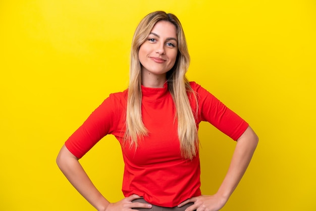 Young Uruguayan woman isolated on yellow background posing with arms at hip and smiling