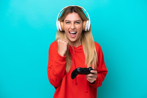 Young Uruguayan woman playing with a video game controller isolated on blue celebrating a victory in winner position