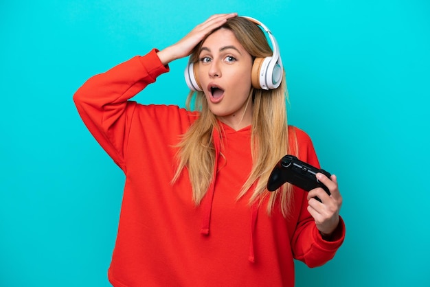 Young Uruguayan woman playing with a video game controller isolated on blue doing surprise gesture while looking to the side