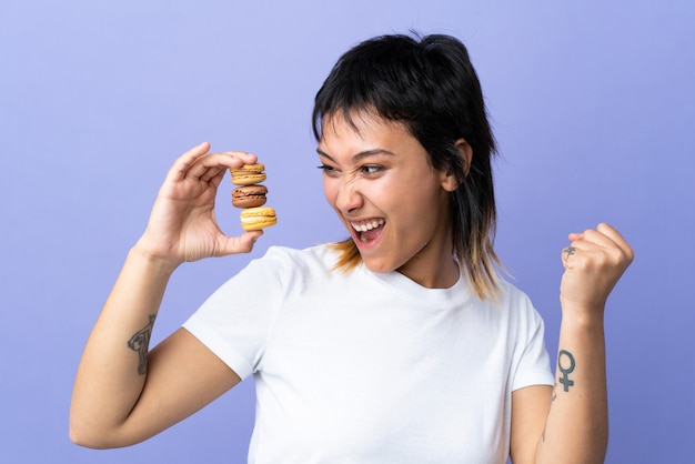 Young Uruguayan woman over purple wall holding colorful French macarons and celebrating a victory