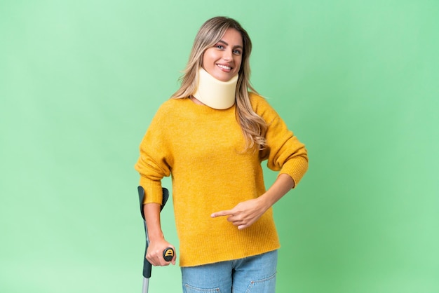 Young Uruguayan woman wearing neck brace and crutches over isolated background and pointing it