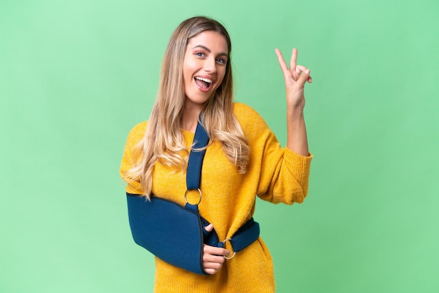 Young Uruguayan woman with broken arm and wearing a sling over isolated background smiling and showing victory sign